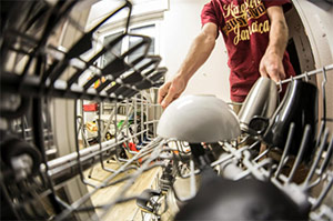 Person loading dishes into a dishwasher, viewed from inside the machine. Plates and cups are visible in the dishwasher rack, and the individual is wearing a red t-shirt.